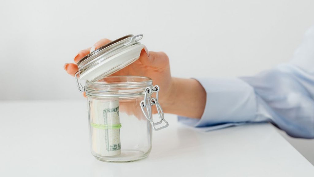 Hand opening a glass jar of rolled up cash savings to show the personal savings allowance